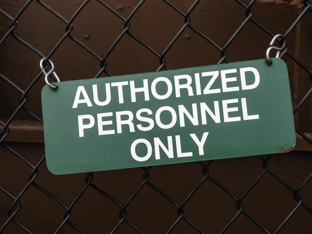 Light green outdoor sign, slightly crooked, on black chain-link fence by dark red steel wall AUTHORIZED PERSONNEL ONLY